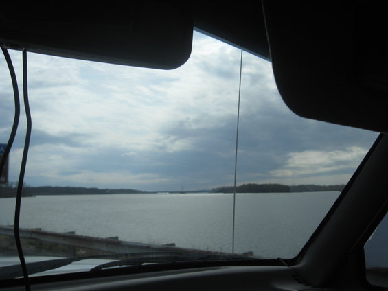 View from inside a vehicle looking out at a large lake or river under a cloudy sky. The dashboard and sun visors are visible