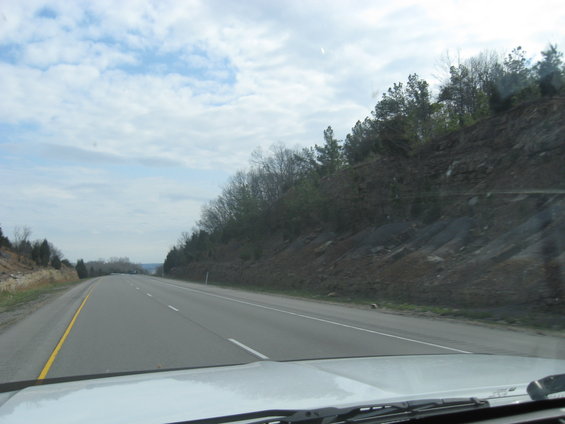 View from inside a vehicle driving on a highway with trees and a rocky embankment on the right