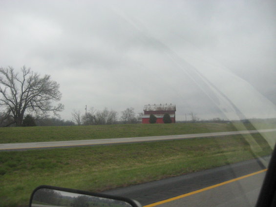 Red barn with white trim sits in a grassy field next to a road under a cloudy sky. Bare tree on left