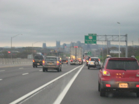 Red SUV and other vehicles on a highway under a cloudy sky, with a city skyline in the distance