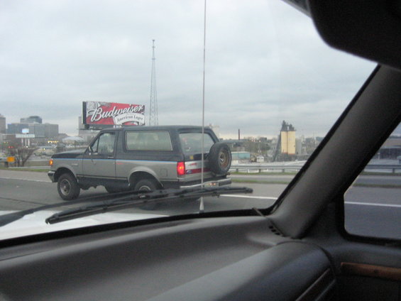 Ford Bronco with spare tire on back driving on highway, Budweiser billboard in background