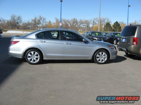 Silver sedan parked on asphalt lot, side view. Visible damage on front fender