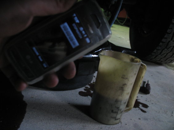 Phone screen shows stopwatch app. Person holds phone over a dirty measuring jug with dark fluid