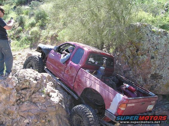 Red Toyota pickup truck with large off-road tires navigating a rocky trail. A person is visible in the driver's seat