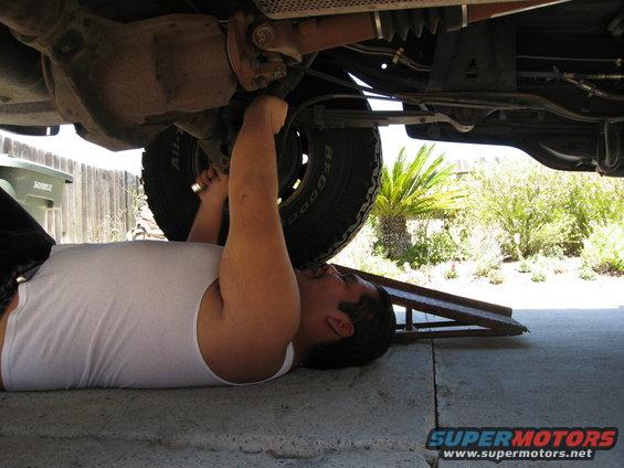 Man in white tank top working on the undercarriage of a vehicle, with a tire visible