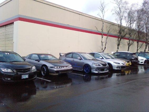 Line of cars parked on a wet lot, including a silver sedan with a spoiler