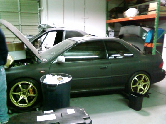 Dark green coupe with gold wheels, hood open, parked next to another car in a garage. Trash can nearby