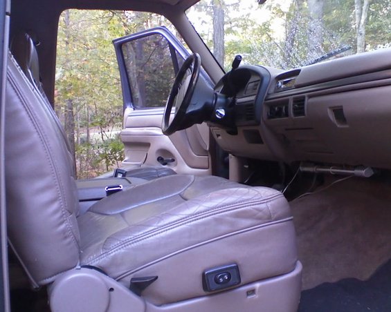 Interior of a 1996 Ford Bronco, showing the driver's seat, dashboard, and steering wheel. Door is open, trees visible outside