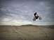 1900 Honda Big Red - A rider on a red dirt bike performs a jump over sand dunes under a cloudy sky.