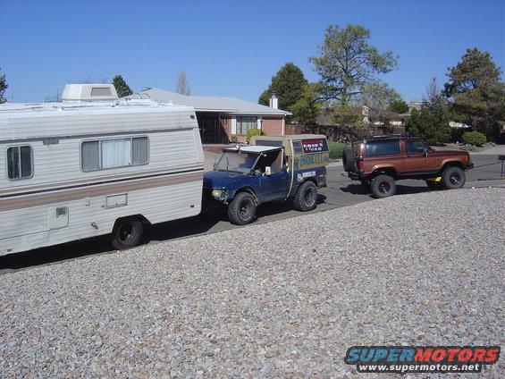 RV trailer towed by blue pickup, brown SUV follows. Text on pickup reads 'BRONCO II'