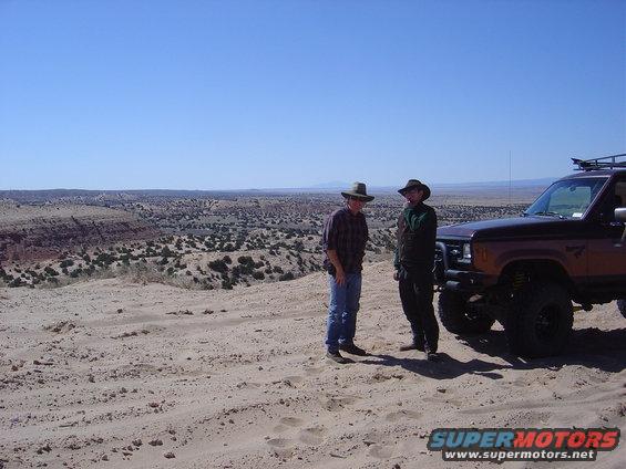 Two guys in cowboy hats stand next to a lifted, dark brown SUV with a roof rack in a sandy, desert area. The SUV has a brush guard