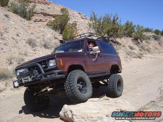 1986 Ford Bronco II with roof rack and bull bar driving over rocks on a dirt trail