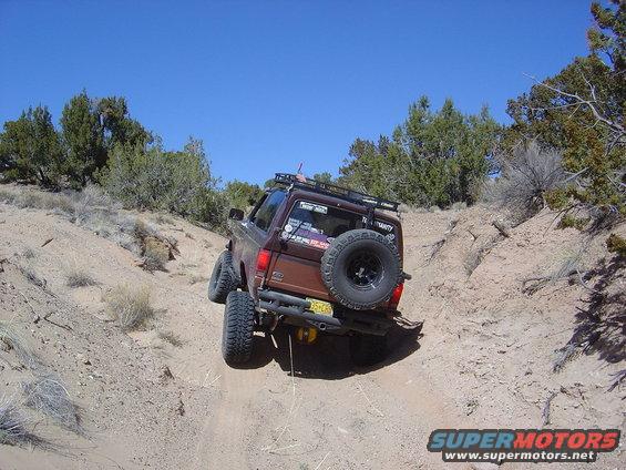 Brown SUV with off-road tires and roof rack driving on a dirt path, spare tire mounted on rear door