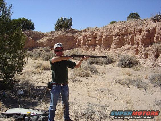 Man in jeans and t-shirt aims a rifle in a desert with rocky hills behind him. Supermotors.net logo in lower right