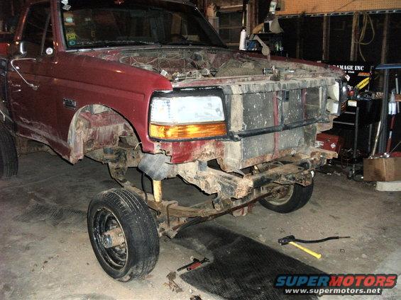 Red Ford F-series truck with front end disassembled, showing exposed engine bay and suspension components