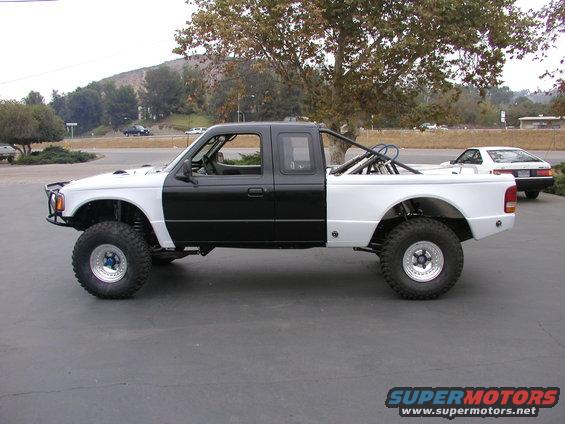 Modified pickup truck with black cab and white bed, large off-road tires, and roll cage. Another car is parked in the background