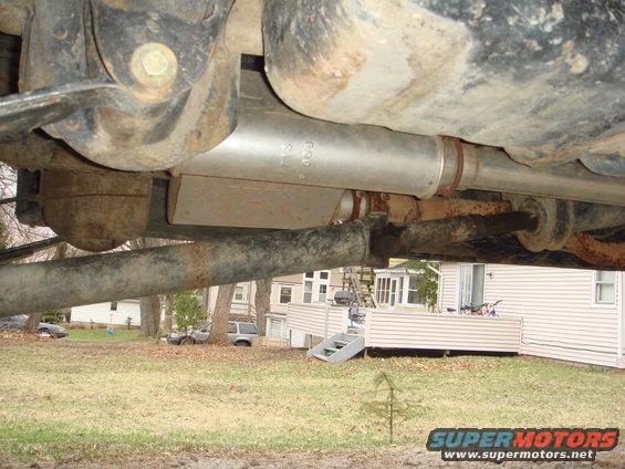 Underside view of a vehicle's exhaust system and suspension components. Houses and other vehicles visible in the background
