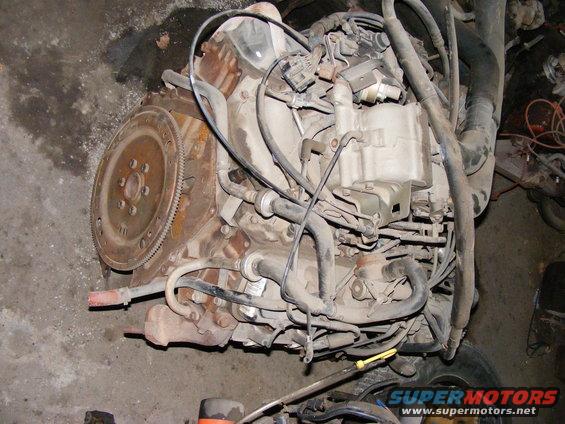 Engine block with flywheel, hoses, and wiring. Rusty and dirty