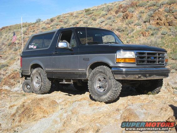 Two-tone Ford Bronco on rocky terrain. American flag on a pole attached to the rear