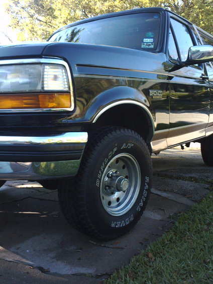 Dark blue Ford Bronco with chrome trim and a tan stripe, parked on a driveway. The front fender and wheel are visible