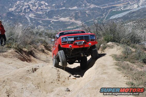 Red SUV with off-road tires climbing a dirt hill. Website address on windshield