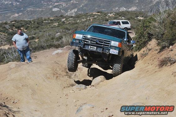 Green Ford Bronco navigating a rocky trail with another Bronco behind it. A man stands to the left