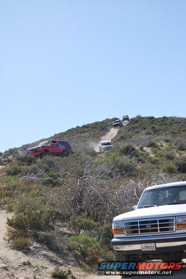 White Ford truck in foreground, other SUVs driving up a dusty hill