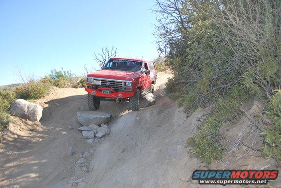 Red Ford Bronco modified for off-roading, climbing over rocks on a dirt trail. Has a winch and aftermarket bumper