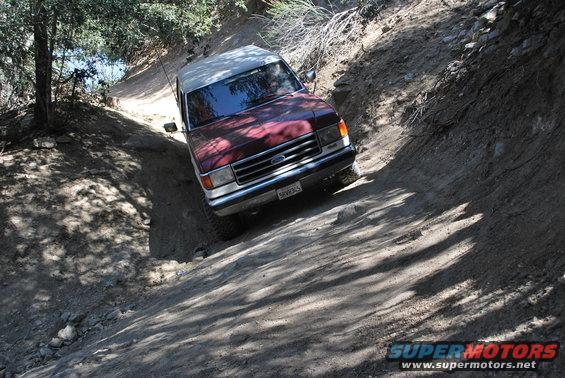 Red and white Ford Bronco navigating a steep, rutted dirt path with trees on the left
