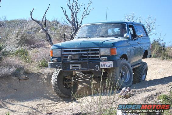 Blue Ford Bronco with large tires driving on a dirt trail with dry brush and trees