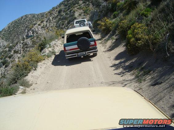 Several SUVs driving up a steep, dusty mountain road. The lead vehicle is a Ford Bronco
