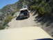 1986 Ford Bronco - A convoy of off-road vehicles navigates a narrow, winding dirt path on a mountainside under a clear blue sky.