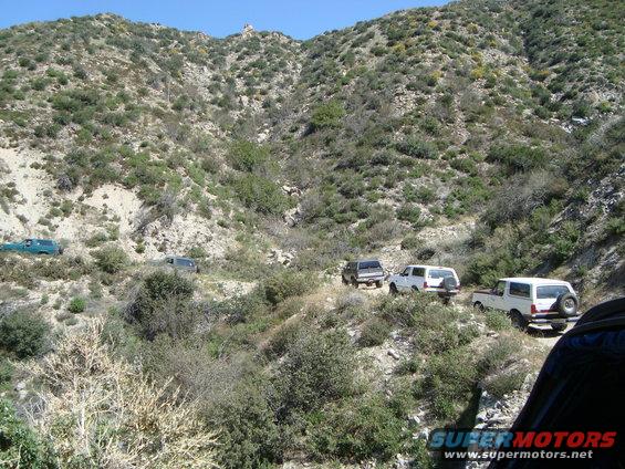 White Ford Bronco with spare tire on rear, white SUV, and dark SUV driving on a dirt road in a canyon