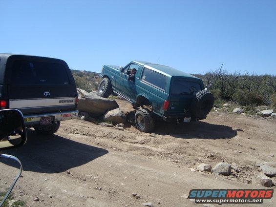 Green 1986 Ford Bronco off-roading over rocks, rear spare tire visible