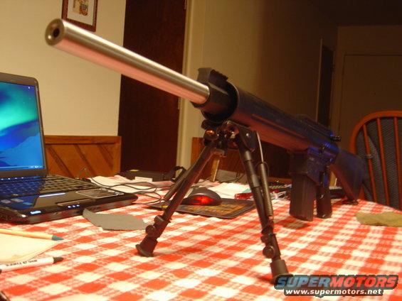 Rifle with bipod on a red and white checkered tablecloth, next to a laptop