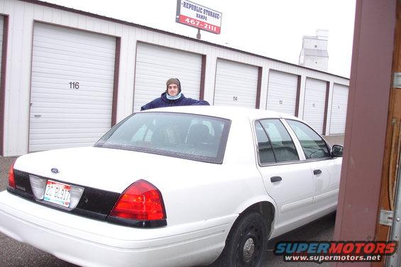 White sedan parked in front of storage units. Person leaning on roof. License plate visible