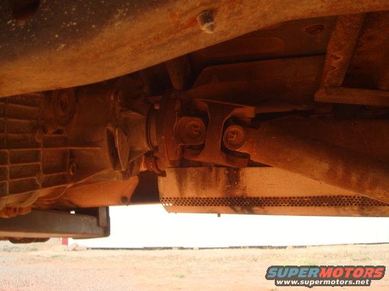 Underside view of a vehicle's transmission and driveshaft, showing rust and dirt