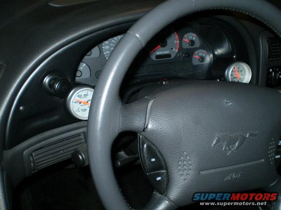 Interior shot of a 2002 Ford Mustang showing the steering wheel with a horse emblem, dashboard gauges, and an aftermarket fuel gauge