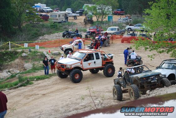 Off-road trucks and ATVs gathered on a dirt track with spectators