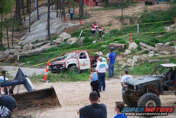 Two off-road trucks are parked on a dirt track with spectators around. One truck is white with a red roll cage, the other is dark colored