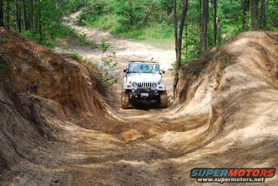 White Jeep Wrangler TJ model driving up a steep dirt trail with trees on either side