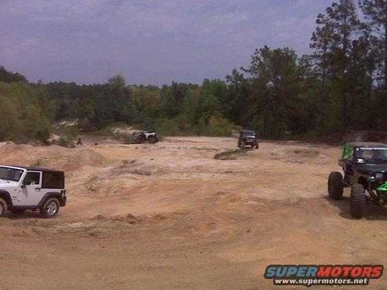 Several Jeeps and off-road vehicles are parked and driving on a sandy, hilly terrain with trees in the background