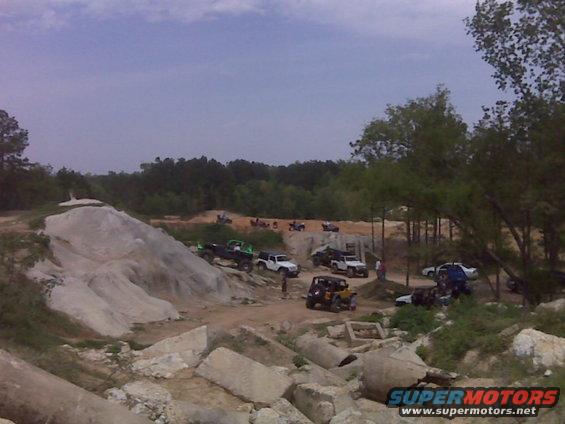 Several off-road vehicles, including Jeeps and trucks, gathered at a dirt park with rocky terrain and trees