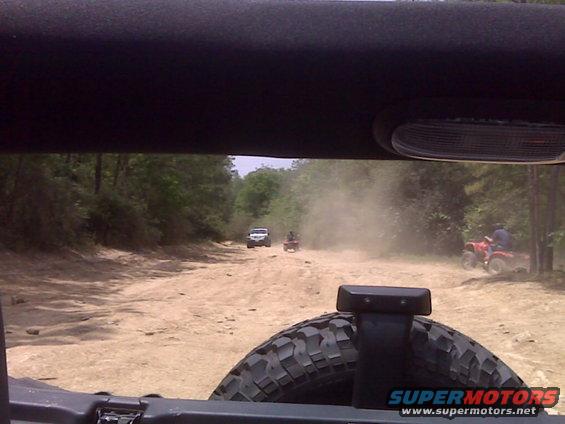 View from inside a vehicle looking out at a dusty dirt road with multiple ATVs and a white SUV