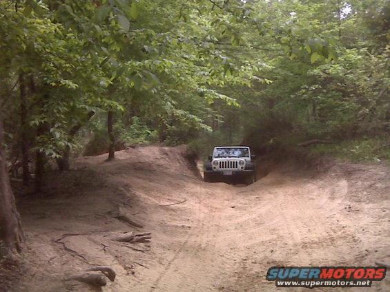White Jeep Wrangler driving up a dirt path in a wooded area. The grille looks like a 2007 model