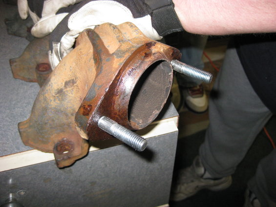 rusted exhaust manifold with two bolts attached, being held by gloved hands