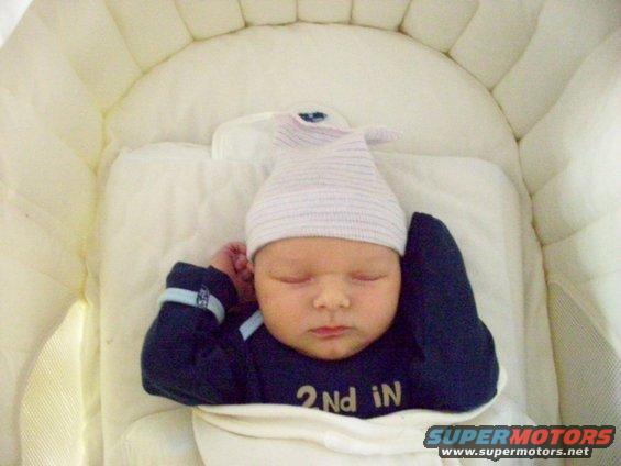 Baby sleeping in a crib wearing a hat and dark blue sleepsuit with '2nd iN' printed on it