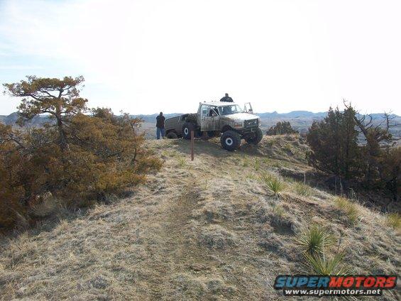 White off-road truck with large tires parked on a grassy hill, two people standing nearby