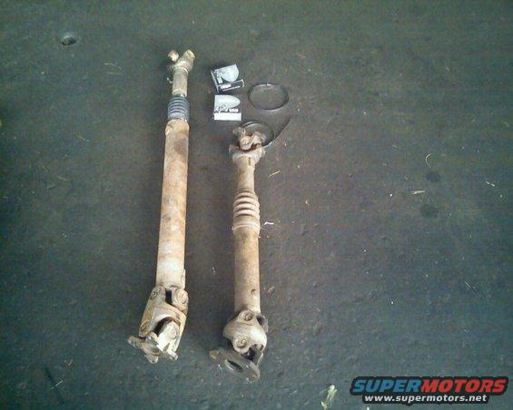 Two rusty driveshafts with U-joints and a spring boot, one longer than the other, on a dark concrete floor