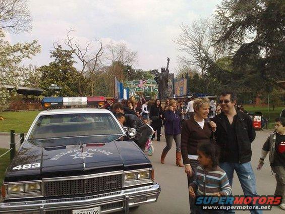 Police car with 'HIGHWAY PATROL' on hood, people in background near a statue